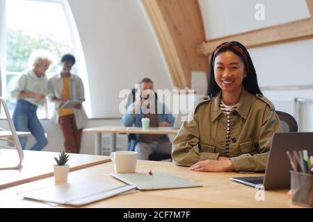 Portrait of Asian young woman smiling at camera while working at her workplace with other people in the background Stock Photo