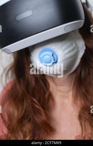 anonymous female with red hair using face mask while experiencing virtual reality using modern headset during quarantine time of coronavirus Stock Photo