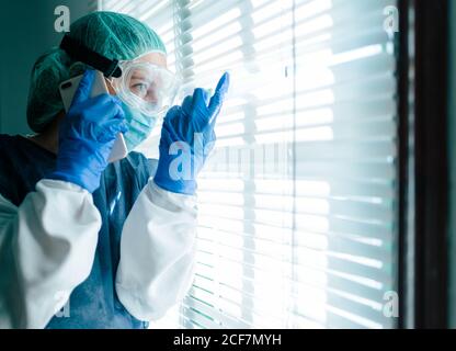 Side view of female medical specialist in protective gown with mask and goggles talking on mobile phone and looking out shuttered window while working in hospital during coronavirus outbreak Stock Photo