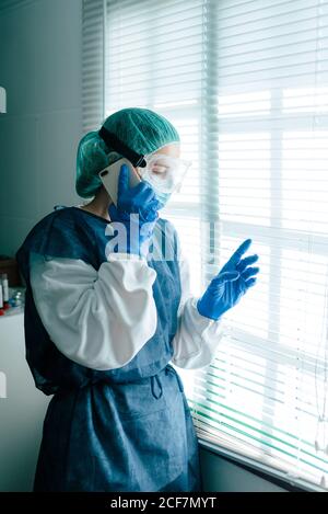 Side view of female medical specialist in protective gown with mask and goggles talking on mobile phone and looking out shuttered window while working in hospital during coronavirus outbreak Stock Photo
