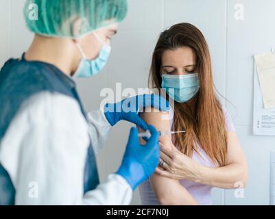 Young medical specialist in protective costume with mask and latex gloves vaccinating female patient in clinic during coronavirus outbreak Stock Photo
