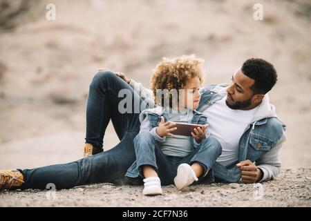 African American bearded casual man lying on sandy ground leaning on elbow and bending leg at knee looking at curly ethnic toddler in denim clothes sitting beside with mobile phone Stock Photo