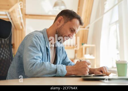 Serious mature businessman sitting at the table and writing he doing paperwork at office Stock Photo