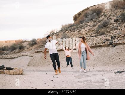 Happy diverse mother father and curly child wearing bright casual clothes strolling on nature holding hands at daytime Stock Photo