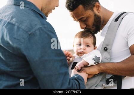 Side view of black casual man holding hand of anonymous male boyfriend and holding little calm baby in grey carrier while strolling on nature at daytime Stock Photo