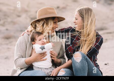 Happy Woman in hat sitting on sandy ground holding little baby in white clothes while female friend supporting touching shoulders in windy weather at daytime Stock Photo