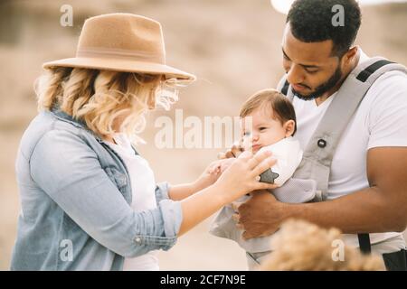 Happy black bearded man carrying little baby and holding hand of blond wife wearing denim shirt jeans and hat while standing on sandy land Stock Photo