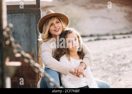 Content Woman with blond hair in hat cuddling daughter in casual clothes while having rest and sitting in old truck body Stock Photo