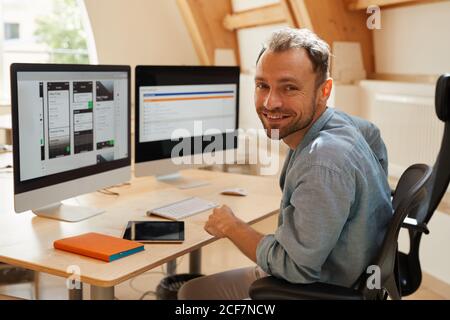 Portrait of programmer smiling at camera while sitting at his workplace with computer monitors and working with software Stock Photo