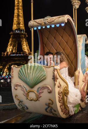 Happily married young man in blue suit and Woman in wedding dress sharing a moment while taking amusement rides in an embrace with Eiffel Tower on background Stock Photo