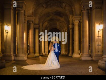 Young newly married couple in wedding suit and gown hugging while standing in rocked arch with Louvre on background at Paris Stock Photo