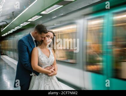 Young bride in white wedding dress standing with closed eyes while groom standing behind and hugging with moving train on blurred background at subway Stock Photo