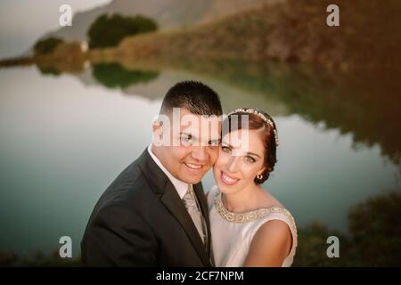 Charming smiling bride and content groom hugging and looking at camera on background of calm crystal lake surrounded by greenery and hills Stock Photo