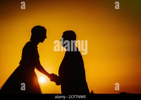 Side view of bright contrast silhouette of amorous man and graceful Woman holding hands in orange light of sunset Stock Photo