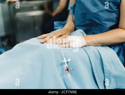 Crop female patient sitting with covered legs and intravenous fluid needle in hand before surgery in operating room Stock Photo
