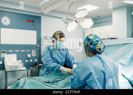 Medics in blue surgical gowns and protective masks holing pipes in body of covered patient lying on bed Stock Photo