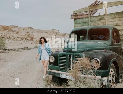 Young Woman in shorts and denim shirt walking by green truck with wooden body and looking away on nature background Stock Photo