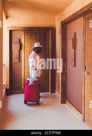 Adult Woman in and casual white clothes with red suitcase closing wooden door in hotel corridor Stock Photo