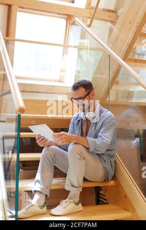 Mature man in eyeglasses sitting on stairs smiling and watching something on digital tablet Stock Photo