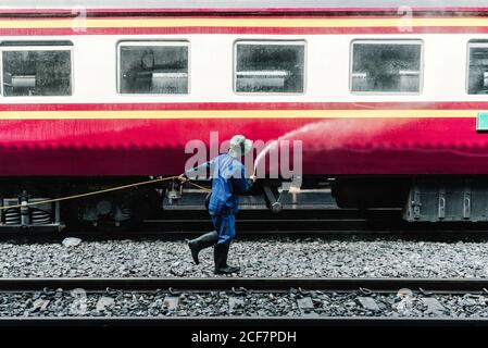 Side view of faceless male worker in blue uniform and hat pouring water from hose in red train on railroad station Stock Photo