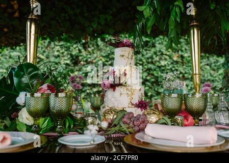 White and gold tiered cake with flowers placed on wedding table with plate and glasses decorated with fruits and flowers against green trees Stock Photo
