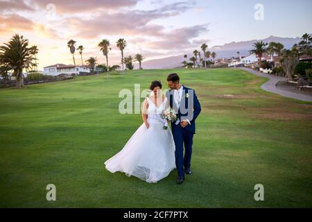 Cheerful groom in elegant suit and bride in white wedding dresses cuddling and walking on lawn while looking at each other during wedding celebration Stock Photo