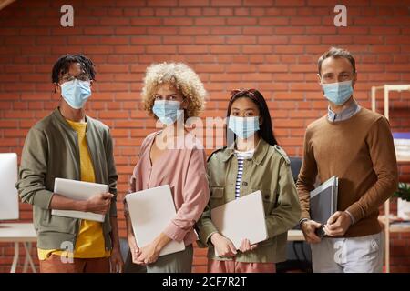 Portrait of group of software developers in protective masks holding their computers and looking at camera while standing at office Stock Photo