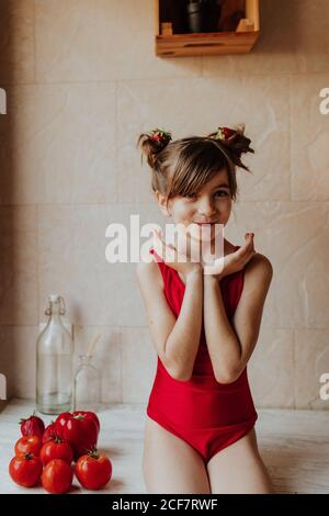 Cute barefoot girl in red bodysuit sitting on counter with tomatoes and red pepper in kitchen Stock Photo