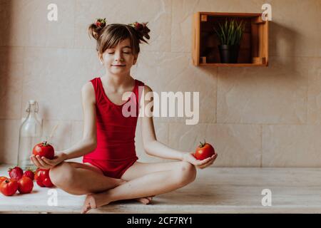 Cute barefoot girl in red bodysuit sitting cross legged on counter and meditating with tomatoes in kitchen Stock Photo