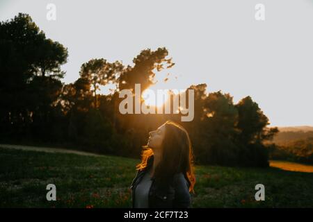 Side view of relaxed young female in black jacket enjoying nature and freedom while walking on beautiful meadow on sunset behind trees