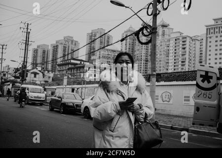 Shanghai, China - 12 December, 2018: Serious adult Chinese female in long warm jacket looking at camera while walking on city street with mobile phone in hands Stock Photo