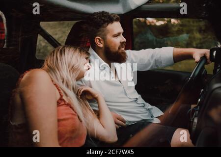 Side view through glass of pleased adult blond Woman in red clothes leaning on shoulder of bearded man in white shirt and black shorts driving car in forest Stock Photo