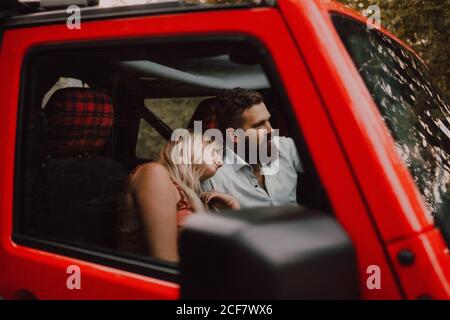 Side view through glass of pleased adult blond Woman in red clothes leaning on shoulder of bearded man in white shirt and black shorts driving car in forest Stock Photo