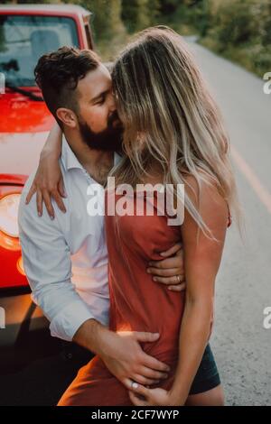 Tender bearded man in white shirt and black shorts hugging smiling Woman in dress while sitting on red car hood on highway near green forest Stock Photo