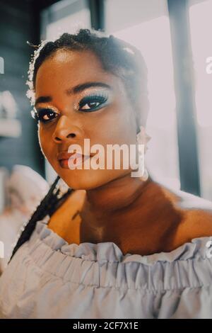 Portrait of backlit beautiful curvy black young Woman with bright make-up in off-shoulder dress standing in cafe on sunset Stock Photo