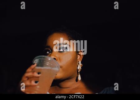 From below portrait of backlit beautiful curvy black young woMan with bright make-up in off-shoulder dress having a cold beverage drink