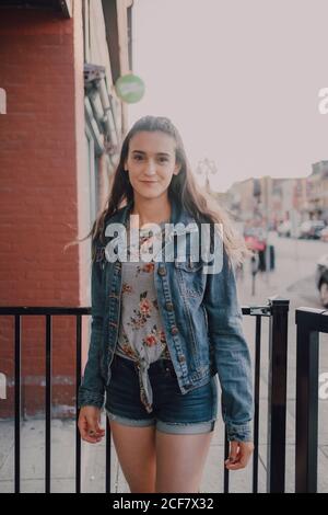 Pretty young young Woman with long hair in casual jeans jacket and shorts standing on street on sunset looking at camera Stock Photo