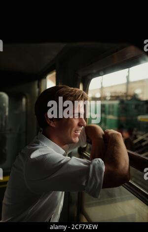 Side view of happy young male in shirt smiling and looking out train window during stop on railway station Stock Photo