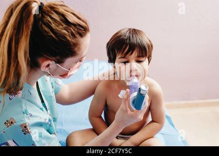 Side view of young female pediatric doctor in medical mask doing inhalation treatment with nebulizer for little boy having respiratory problem in hospital Stock Photo