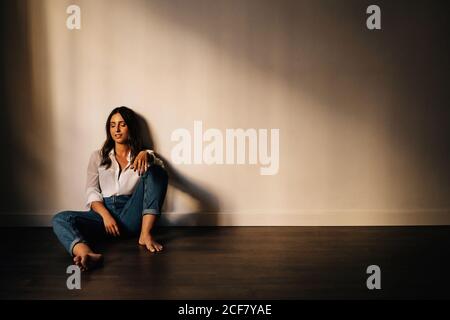 Gorgeous barefoot brunette in white shirt and blue jeans smiling with closed eyes while sitting on floor near wall in empty room Stock Photo