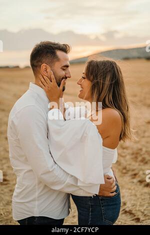 Side view of happy man and Woman hugging and looking at each other while standing against dry meadow and sunset sky Stock Photo