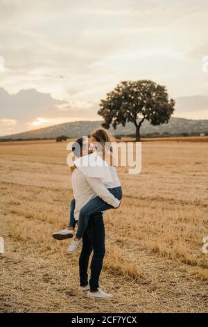Side view of happy man hugging and carrying girlfriend while standing on dry grass of meadow against sundown sky and tree Stock Photo