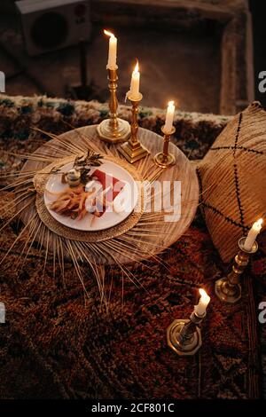 From above composition in Arabic style with small round table with burning candles and herbs on plate placed on carpet near cozy pillow Stock Photo