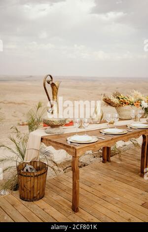 Long wooden table decorated with flowers and candles and served with white plates on traditional Arabic set with sandy dunes in background Stock Photo