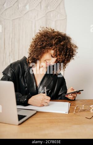 Positive retro businessWoman with curly hair smiling and writing in notebook while sitting at table and using smartphone in office Stock Photo
