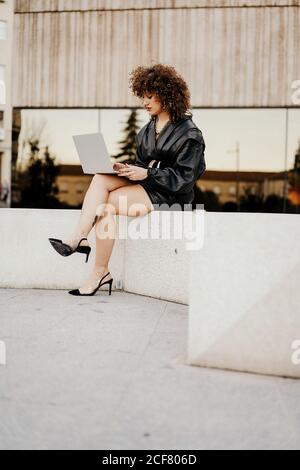 businessWoman in black leather outfit sitting on border and browsing data on laptop while working on remote project on city street Stock Photo