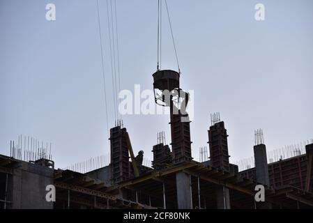 Crane lifting a concrete bucket on sunset background. Silhouettes of builders pouring cement mix for formworks at a construction site. Soft focus, pos Stock Photo