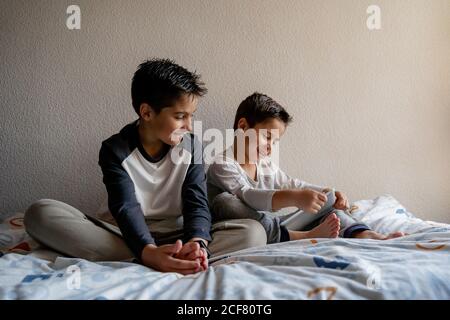 Smiling brothers in sleepwear sitting on cozy bed and watching movie together while entertaining at home Stock Photo
