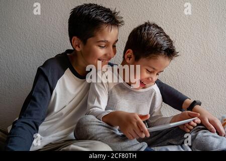 Smiling brothers in sleepwear sitting on cozy bed and watching movie together while entertaining at home Stock Photo