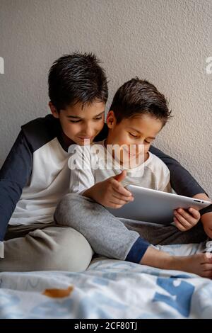 Smiling brothers in sleepwear sitting on cozy bed and watching movie together while entertaining at home Stock Photo